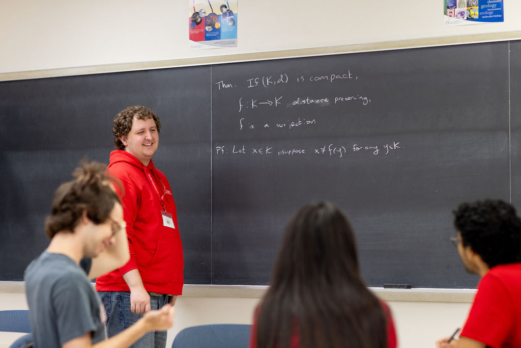 Students collaborate on math problems in a small-group tutoring session, with one student working at a chalkboard while others follow along from a table.